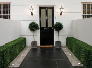 Elegant front entrance featuring Pavestone Black Kadapha limestone paving laid in a linear pattern, bordered by grey gravel strips with recessed lights, neatly trimmed hedges, and topiary trees flanking a black front door