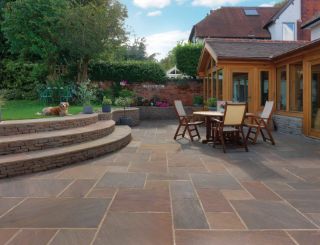 Elegant garden patio featuring Pavestone Calibrated Sandstone in Modak, displaying warm tones of terracotta, rose, and muted plum. The outdoor space includes curved stone steps, a wooden dining set, and landscaped borders, with a spaniel resting on the raised lawn beside a vintage bistro set