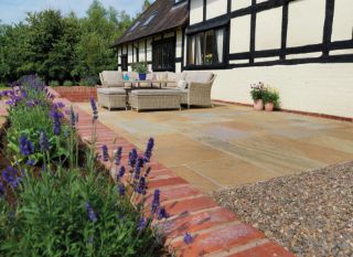 A landscaped patio featuring Pavestone calibrated sandstone slabs in warm, earthy tones, set beside a traditional half-timbered house with wicker outdoor furniture and blooming purple lavender in the foreground