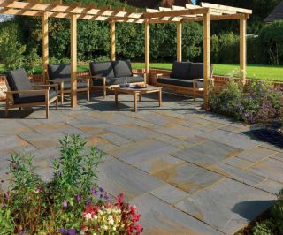 Stylish garden patio paved with Pavestone Calibrated Sandstone in Blue Blend, featuring cool slate-grey tones with hints of bronze. The outdoor space includes modern wooden furniture under a timber pergola, surrounded by lush green lawn, flower borders, and a red brick wall for a relaxed contemporary setting