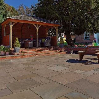 Spacious garden patio paved with Pavestone Calibrated Sandstone in Willow Blend, featuring subtle tones of grey, beige, and olive. The patio includes a timber gazebo with rattan seating, a brick retaining wall with potted flowers, and a wooden picnic bench, all set in a traditional English garden
