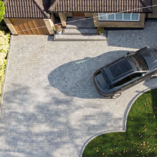 Aerial view of a modern residential driveway paved with Brett Alpha 50mm block paving in Silver Haze. The driveway features a neat curved edge along a well-maintained lawn and a charcoal border, with a parked black SUV in front of a brick house with a tiled roof