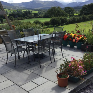 A contemporary garden patio featuring Pavestone Black Kadapha limestone slabs laid in a mixed-size pattern, bordered by lush greenery and outdoor furniture