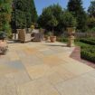 Spacious garden patio featuring Pavestone calibrated limestone slabs in warm golden and grey tones, laid in a mixed-size pattern. The patio is furnished with wicker dining furniture and surrounded by terracotta pots, manicured hedges, and mature trees