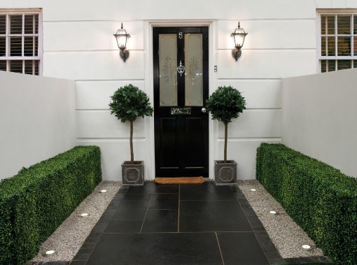 Elegant entranceway featuring Pavestone Black Kadapha limestone paving laid in a clean linear pattern, bordered by gravel strips with recessed uplighting and neatly clipped hedges. Two topiary trees in square planters frame a black front door with traditional lantern wall lights