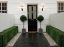 Elegant entranceway featuring Pavestone Black Kadapha limestone paving laid in a clean linear pattern, bordered by gravel strips with recessed uplighting and neatly clipped hedges. Two topiary trees in square planters frame a black front door with traditional lantern wall lights