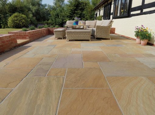 Spacious countryside patio laid with Pavestone Calibrated Sandstone in Rustic Blend, showcasing warm tones of buff, amber, and soft plum. The patio features a wicker outdoor seating set, red brick retaining walls, and potted flowers, all set against a traditional timber-frame cottage