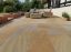 Spacious countryside patio laid with Pavestone Calibrated Sandstone in Rustic Blend, showcasing warm tones of buff, amber, and soft plum. The patio features a wicker outdoor seating set, red brick retaining walls, and potted flowers, all set against a traditional timber-frame cottage