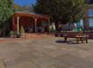 Spacious garden patio paved with Pavestone Calibrated Sandstone in Willow Blend, featuring subtle tones of grey, beige, and olive. The patio includes a timber gazebo with rattan seating, a brick retaining wall with potted flowers, and a wooden picnic bench, all set in a traditional English garden