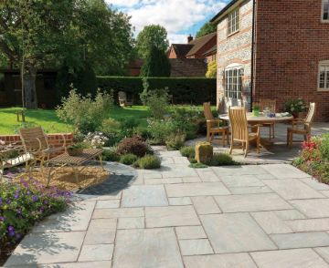 Charming cottage garden patio paved with Pavestone Calibrated Sandstone in Light Grey, featuring soft silver and pale grey tones. The outdoor space includes wooden dining furniture, curved bench seating, flower borders, and landscaped planting, creating a tranquil and inviting setting