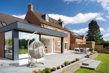 Modern garden patio featuring Pavestone Classic Porcelain Paving Tiles in a light grey finish. The spacious outdoor area includes a hanging egg chair, a dining set, and raised flower beds with stone walling, creating a stylish and contemporary space against a backdrop of a red brick home and clear blue sky
