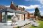 Modern garden patio featuring Pavestone Classic Porcelain Paving Tiles in a light grey finish. The spacious outdoor area includes a hanging egg chair, a dining set, and raised flower beds with stone walling, creating a stylish and contemporary space against a backdrop of a red brick home and clear blue sky