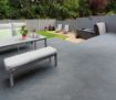 Contemporary garden setting featuring dark grey Pavestone Classic Porcelain Paving Tiles. The patio includes a modern grey dining bench and table with soft cushions, overlooking a lawn area with sun loungers and a hot tub, enclosed by a light grey timber fence and surrounded by lush greenery