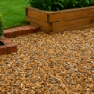 Close-up of a landscaped garden corner featuring gold flint gravel in natural amber, beige, and grey tones, bordered by red brick edging, a wooden planter, and green shrubs under soft daylight