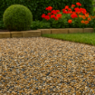 Close-up of a landscaped garden path covered in small 20mm Newbury Style flint gravel, with tones of buff, grey, and amber. The path is bordered by stone edging, with neatly trimmed topiary and vibrant red flowers in the background, under soft daylight.