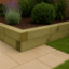Raised garden bed constructed from pressure-treated timber sleepers stacked neatly on a sandstone patio, surrounded by grass and planted with lavender, boxwood, and leafy shrubs in a landscaped setting