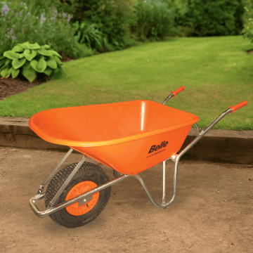 Bright orange Belle Warrior wheelbarrow positioned on a compacted earth surface in a well-maintained garden, with lush grass, green shrubs, and a timber border in the background