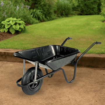 Black Walsall HD wheelbarrow with 85L pressed steel pan and pneumatic tyre, positioned on a compacted soil surface in a landscaped garden with lush grass and green shrubs in the background