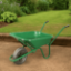 Green Walsall HD wheelbarrow with a 90L pressed steel pan and pneumatic tyre, placed on a compacted soil surface in a landscaped garden with lush grass and dense green foliage in the background