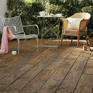 Charming garden patio scene featuring Millboard Weathered Oak decking in a rich brown finish, styled with vintage-style metal and wicker chairs, and a café table, set against lush greenery for a cosy, rustic outdoor retreat