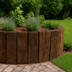 A raised circular garden bed made from vertically placed reclaimed timber sleepers, set on a stone-paved patio with steps to the left. The bed is filled with lush green plants and lavender under bright daylight, creating a rustic garden feature.